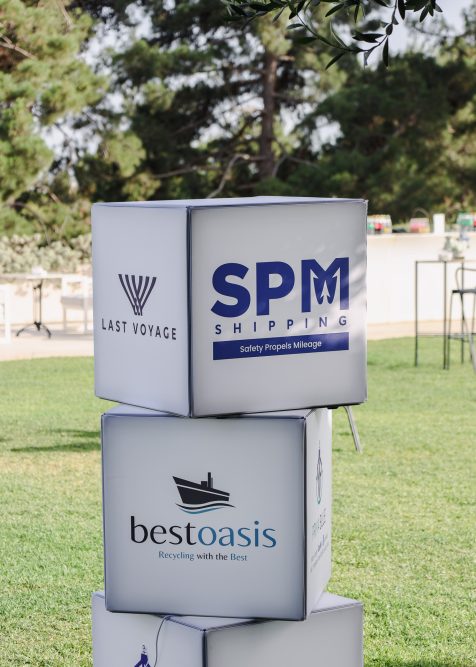 Stacked cubes displaying logos of 'Last Voyage,' 'SPM Shipping,' and 'Best Oasis' on a grassy lawn at the Panhellenic Yachting Congress, with trees in the background