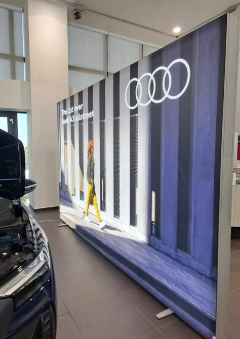 Backdrop display in an Audi showroom featuring the promotion of Audi A3 Allstreet, with an image of a person walking by a modern building