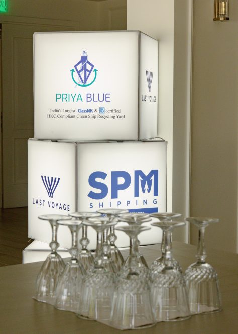 Illuminated cubes displaying logos of 'Priya Blue,' 'Last Voyage,' and 'SPM Shipping' with wine glasses in the foreground at the Panhellenic Yachting Congress.